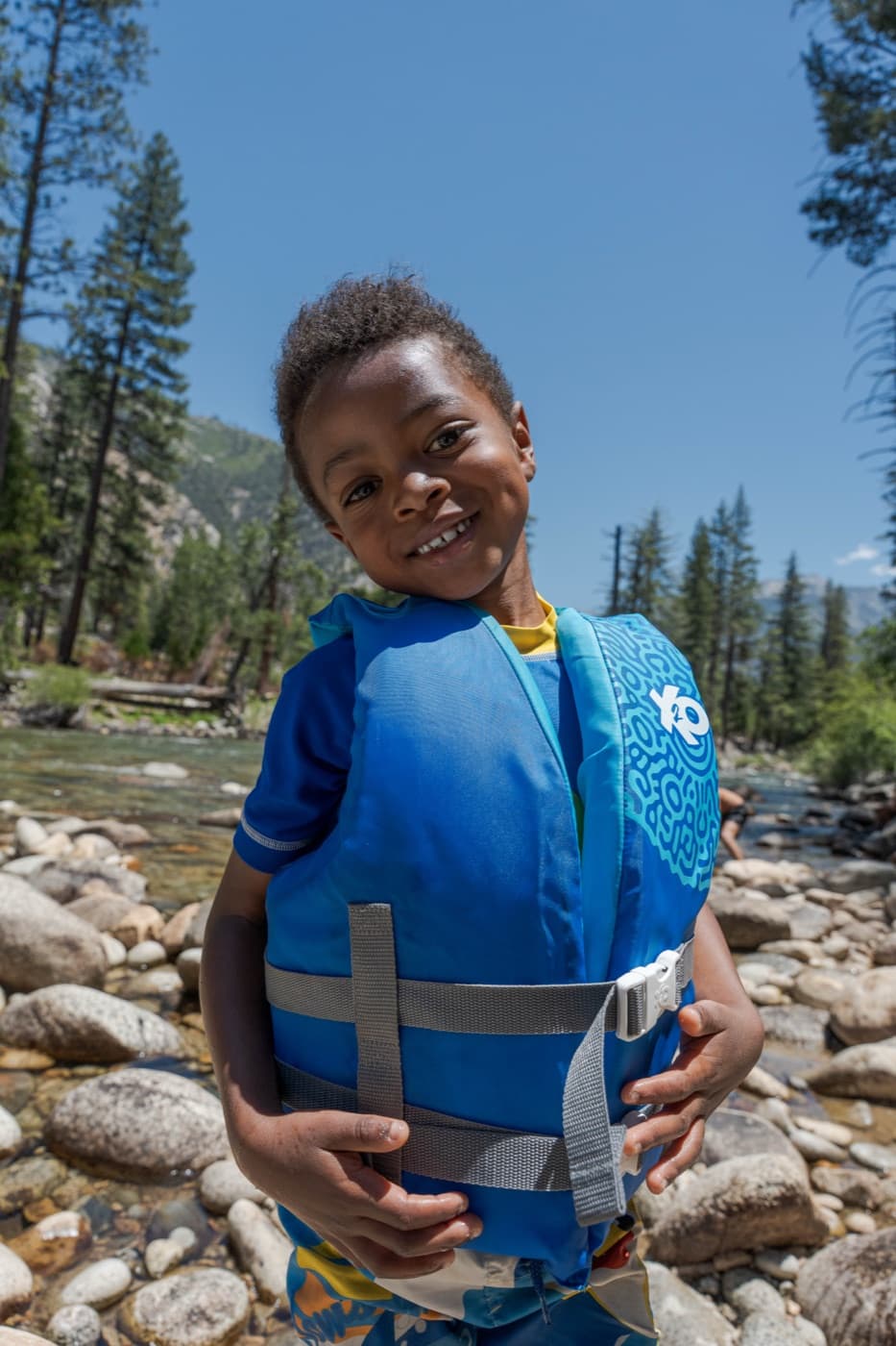 A young boy in a blue life vest beams at the camera on the riverbank at Kings Canyon
