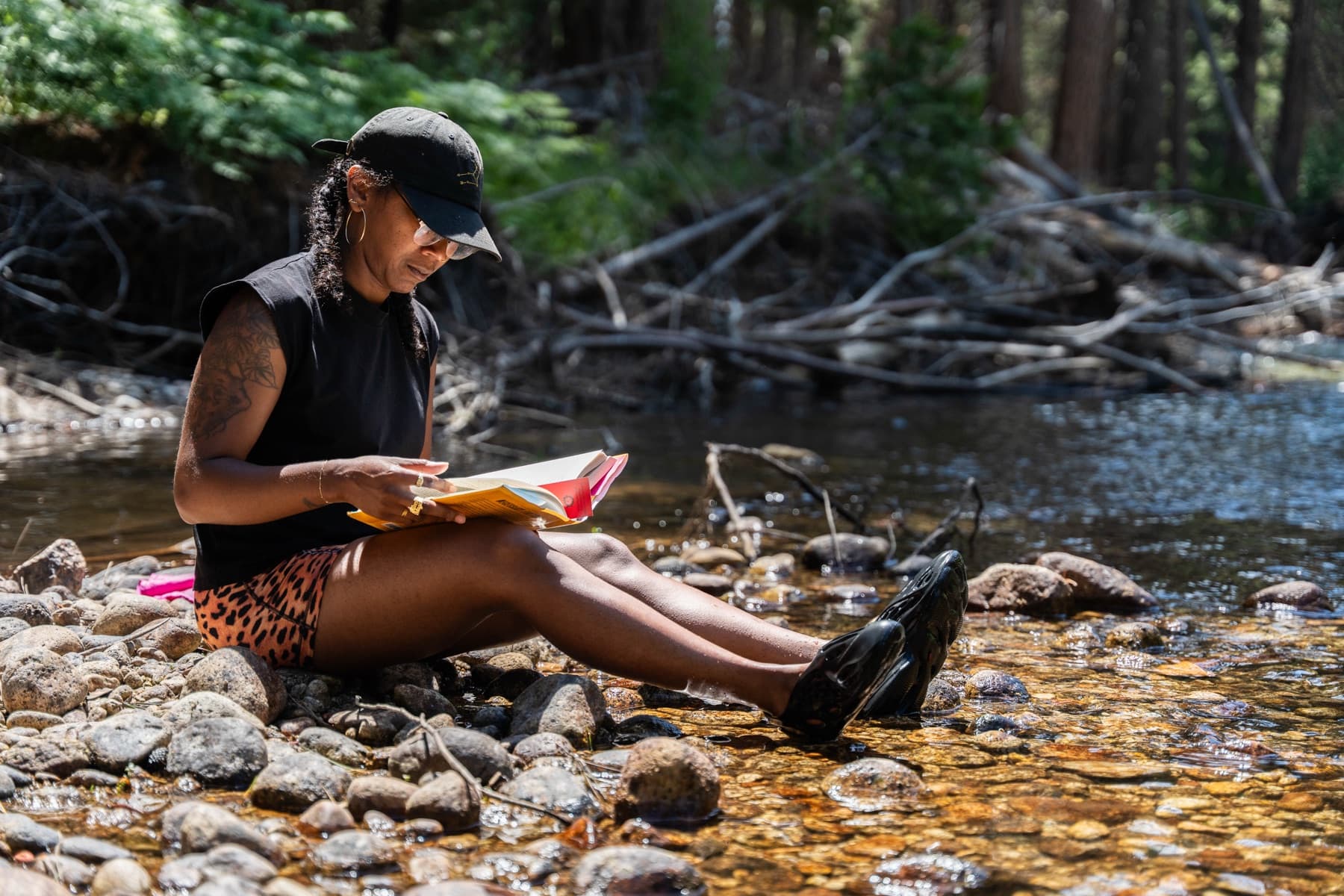 A woman sits reading a book on the rocky bank with her feet in the stream