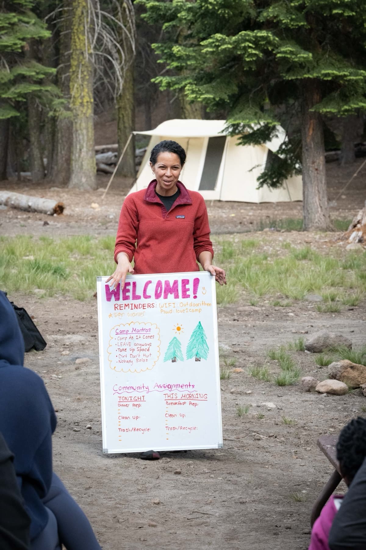 An Outdoorithm host holds a hand-lettered welcome board listing the five mantras and the camp job assignments, in front of canvas wall tents