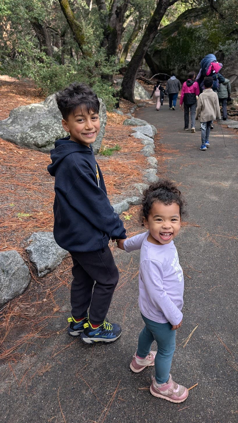 Two children, an older boy and younger girl, smiling outdoors on a hiking trail